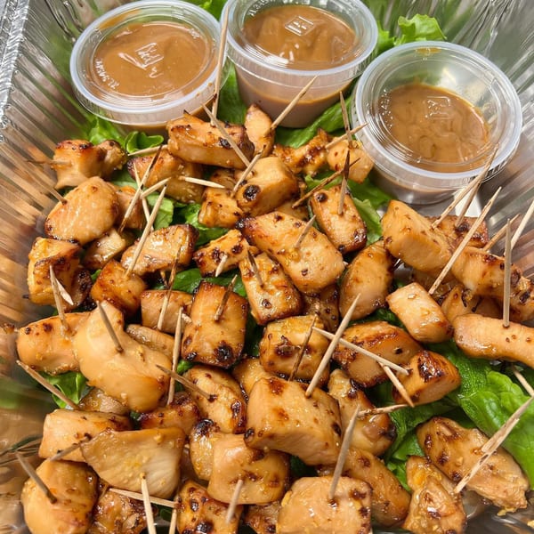 Grilled chicken bites with toothpicks and three small cups of peanut sauce in a foil container.